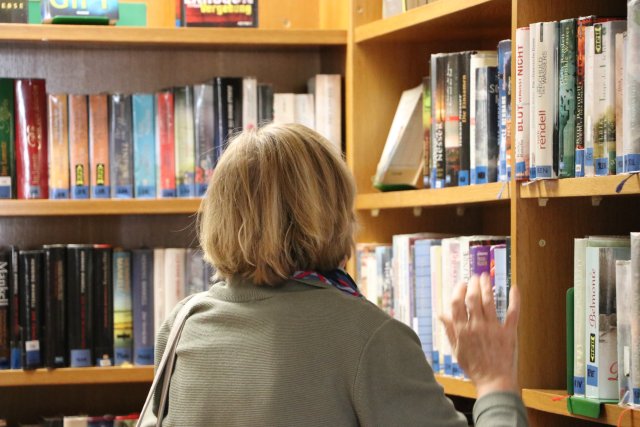 Das Bild zeigt eine Frau in einer Öffentlichen Bibliothek. Sie steht mit dem Rücken zum Betrachter und wählt gerade ein Buch aus.